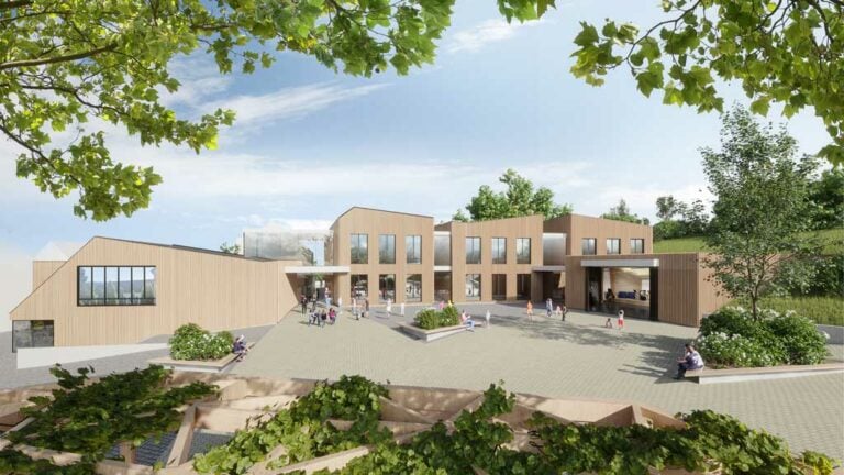 Blick vom Dorfplatz auf die holzverkleideten Kuben der Schule und den Musiksaal des Schulcampus in Bettborn, Kreation der BALLINIPITT Stadtplaner.